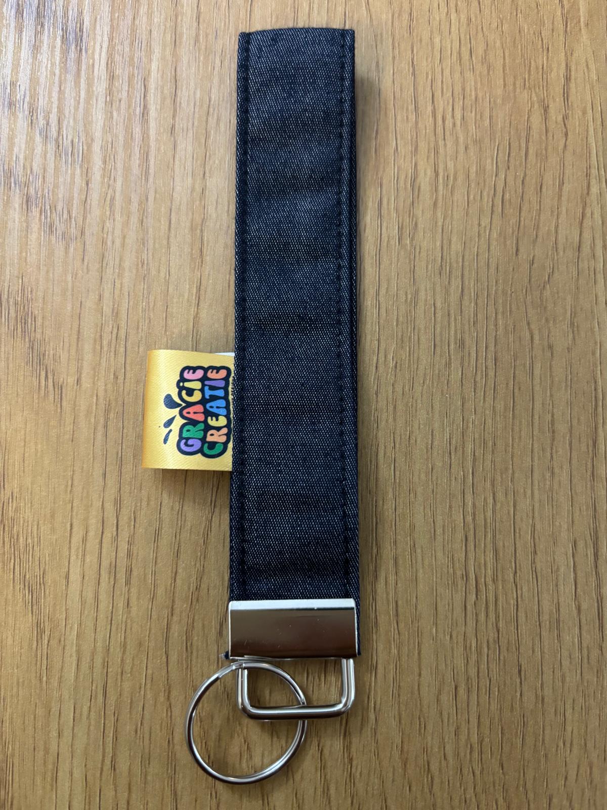 Denim keychain strap with metal clip on a wooden surface, featuring a colorful brand label with metal hardware