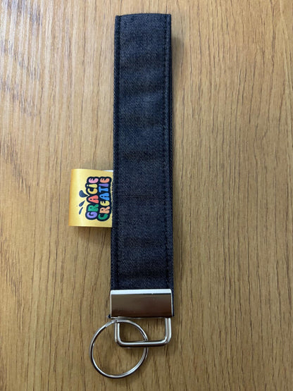 Denim keychain strap with metal clip on a wooden surface, featuring a colorful brand label with metal hardware