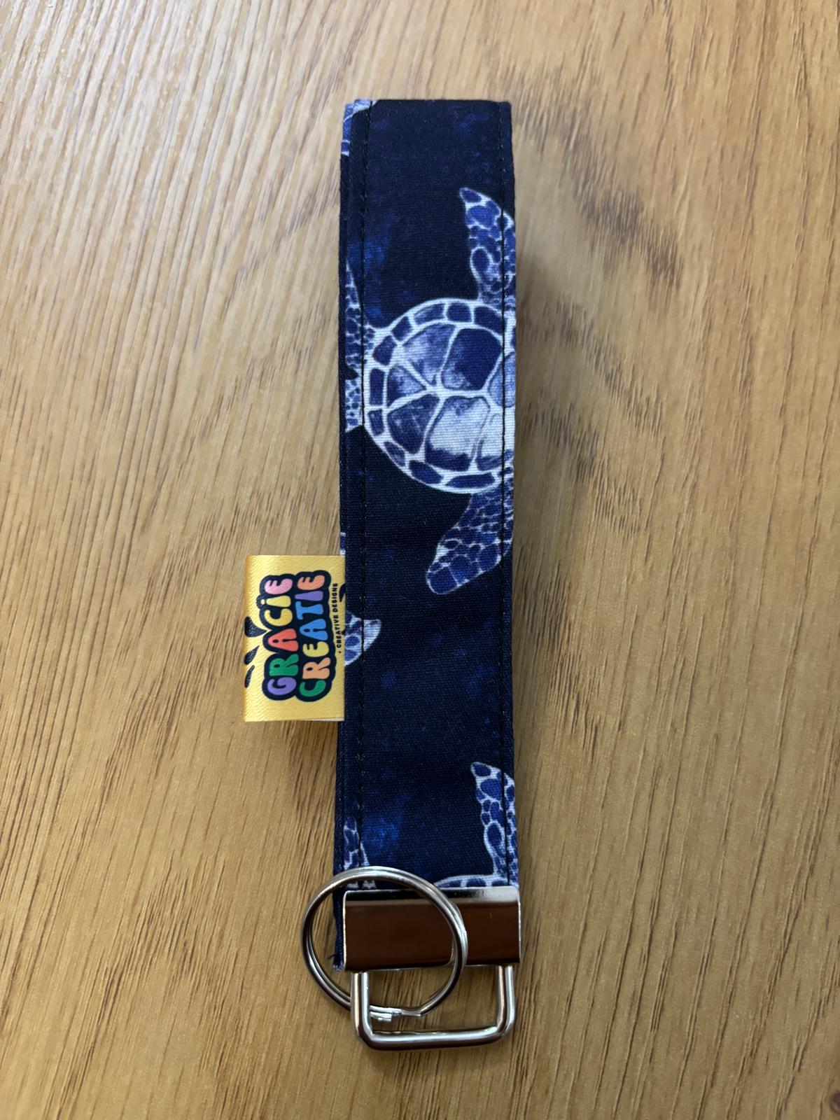 Navy blue key fob with turtle design on a wooden surface, featuring a Gracie Creative tag with metal hardware